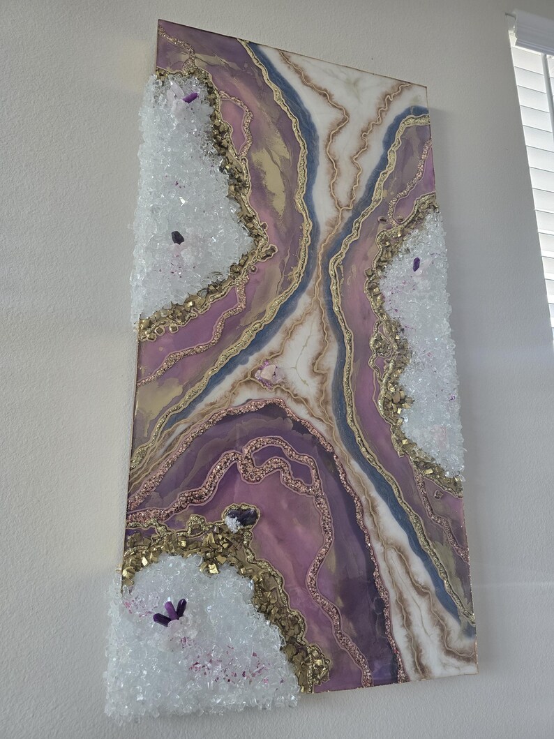 Może przedstawiać: Abstrakcyjna grafika ścienna z motywem geody w odcieniach fioletu, bieli, złota i błękitu. Grafika ma teksturowaną powierzchnię z kryształowymi zdobieniami i złotymi brokatowymi akcentami. Prostokątny element jest zamontowany na białej ścianie.