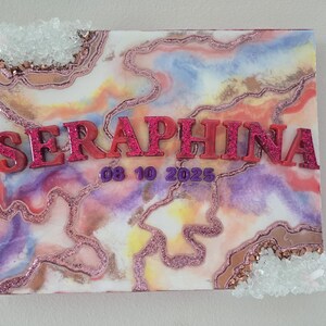 Puede incluir: Un lienzo decorativo con un dise&ntilde;o abstracto colorido en tonos rosa, morado y dorado. El nombre "SERAPHINA" se muestra en letras rosas brillantes en relieve, junto con la fecha "08 10 2025". Acentos de cristal transparente adornan los bordes.
