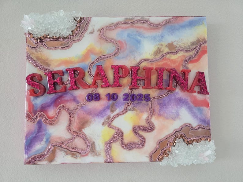Puede incluir: Un lienzo decorativo con un dise&ntilde;o abstracto colorido en tonos rosa, morado y dorado. El nombre "SERAPHINA" se muestra en letras rosas brillantes en relieve, junto con la fecha "08 10 2025". Acentos de cristal transparente adornan los bordes.