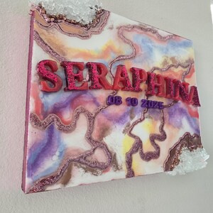 Puede incluir: Una pieza de arte mural decorativa con el nombre "SERAPHINA" en letras rojas con purpurina. La obra presenta un dise&ntilde;o abstracto colorido con tonos rosas, morados y marrones, acentuado con purpurina y adornos de cristal. Tambi&eacute;n es visible la fecha "08 10 2025".