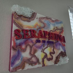 Puede incluir: Una obra de arte decorativa sobre lienzo con el nombre "SERAPHINA" en letras rojas y brillantes. La obra presenta un dise&ntilde;o abstracto colorido con tonos rosas, morados y amarillos, y est&aacute; acentuada con adornos de cristal.