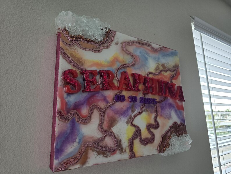 Puede incluir: Una obra de arte decorativa sobre lienzo con el nombre "SERAPHINA" en letras rojas y brillantes. La obra presenta un dise&ntilde;o abstracto colorido con tonos rosas, morados y amarillos, y est&aacute; acentuada con adornos de cristal.