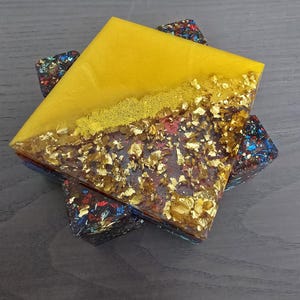 Pode incluir: Uma base de resina quadrada com uma metade superior amarela e uma metade inferior com flocos de folha de ouro. A base assenta sobre tr&ecirc;s bases menores com um design colorido e salpicado. A base tem aproximadamente 10 cm quadrados.