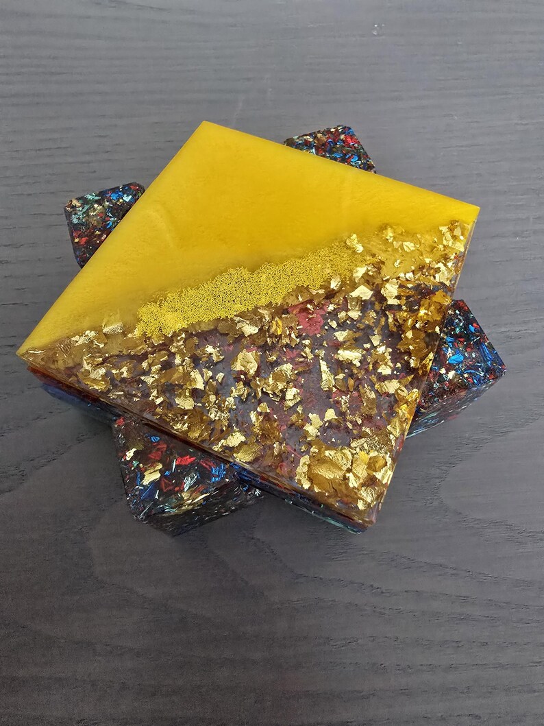 Pode incluir: Uma base de resina quadrada com uma metade superior amarela e uma metade inferior com flocos de folha de ouro. A base assenta sobre tr&ecirc;s bases menores com um design colorido e salpicado. A base tem aproximadamente 10 cm quadrados.
