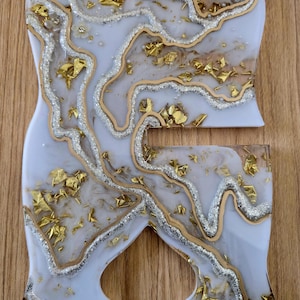 K&ouml;nnte beinhalten: Zweiteiliges Kunstset aus Harz in Wei&szlig;, Gold und Silber. Die Teile haben einen Marmoreffekt mit Blattgold und Glitzerakzenten. Das Set befindet sich auf einer Holzoberfl&auml;che.