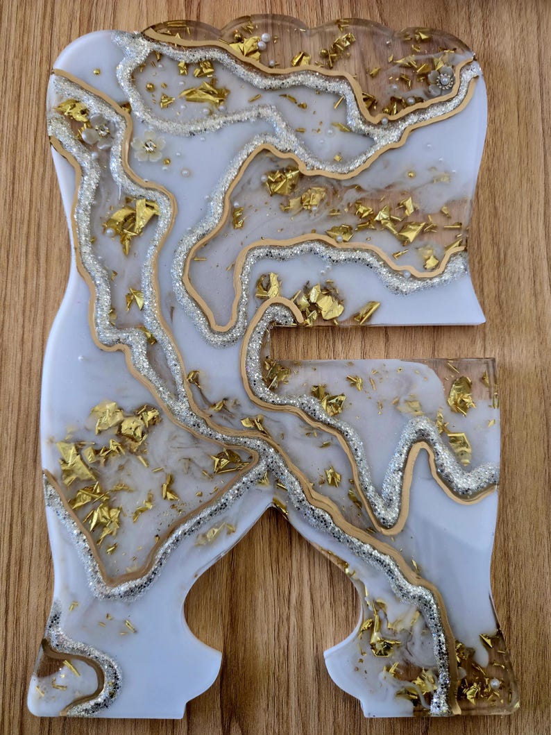 K&ouml;nnte beinhalten: Zweiteiliges Kunstset aus Harz in Wei&szlig;, Gold und Silber. Die Teile haben einen Marmoreffekt mit Blattgold und Glitzerakzenten. Das Set befindet sich auf einer Holzoberfl&auml;che.