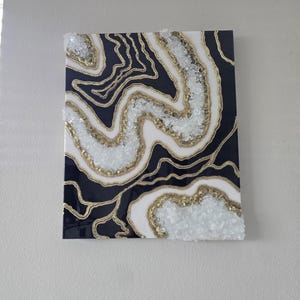 May include: Abstract wall art with a geode design. The artwork has a dark blue background with white and gold accents. The design incorporates a layered effect with crystal-like embellishments, creating a textured appearance. The piece is mounted on a square canvas.