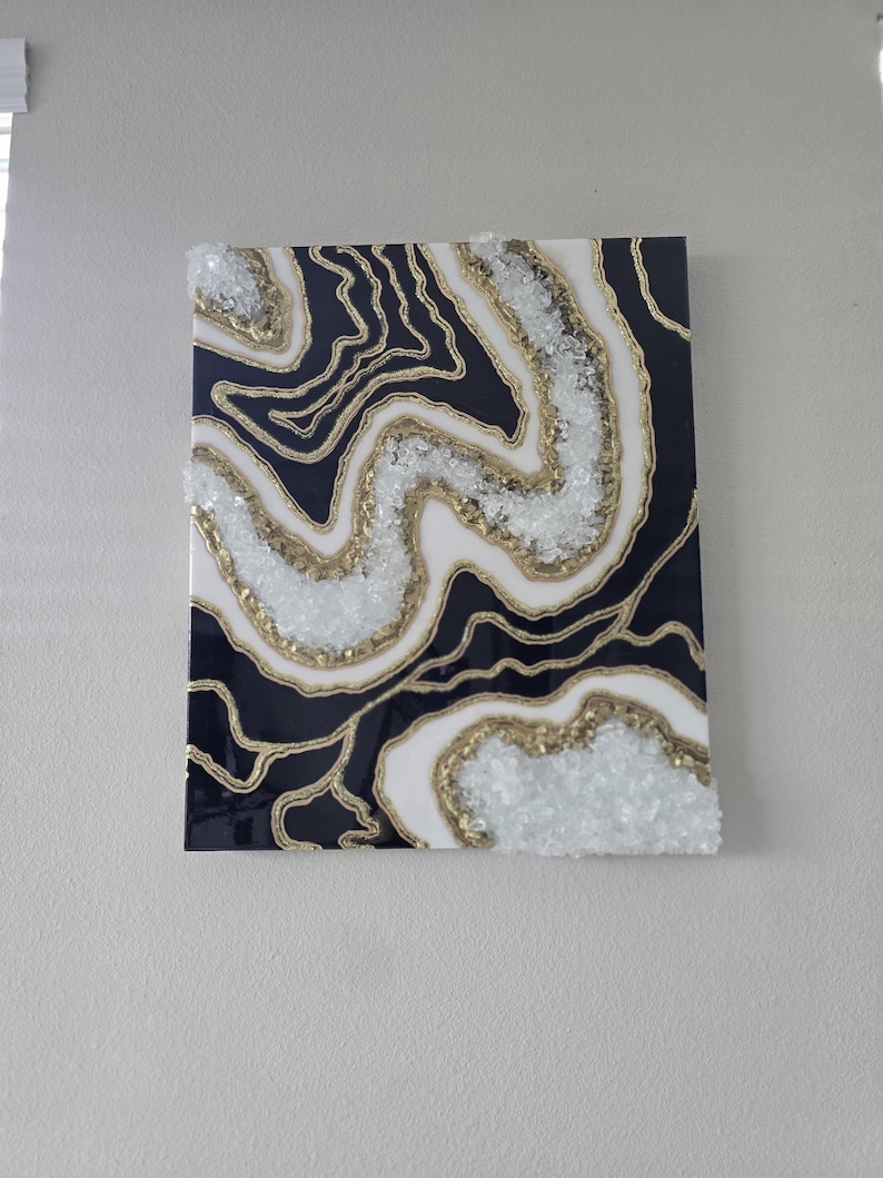 May include: Abstract wall art with a geode design. The artwork has a dark blue background with white and gold accents. The design incorporates a layered effect with crystal-like embellishments, creating a textured appearance. The piece is mounted on a square canvas.