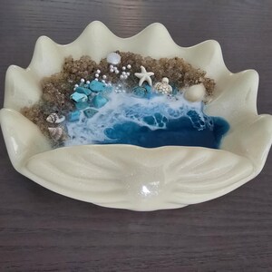 May include: An off-white clam shell-shaped dish with a miniature ocean scene. The scene features a blue resin ocean, sand, turquoise stones, seashells, a starfish, and tiny turtle figurines. The dish has a glossy finish.