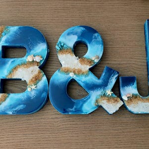 May include: The letters B and J and an ampersand symbol crafted from resin, featuring a blue and white ocean wave design with embedded sand, small seashells, and tiny starfish accents along the edges of each character.
