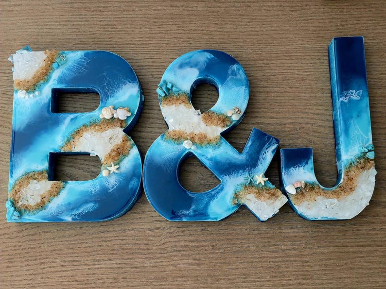 May include: The letters B and J and an ampersand symbol crafted from resin, featuring a blue and white ocean wave design with embedded sand, small seashells, and tiny starfish accents along the edges of each character.
