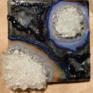May include: Square resin art piece with a galaxy theme. The artwork features a dark background with swirls of black, silver, and blue. Clear, crystalline formations and iridescent glitter add texture and sparkle. The piece has a geode-like appearance.