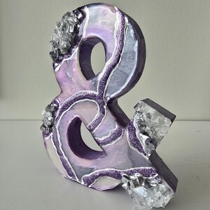 May include: A decorative ampersand sculpture in shades of purple, white, and silver. The sculpture features a glittery, marbled design with crystal-like accents. The ampersand is approximately 8 inches tall.