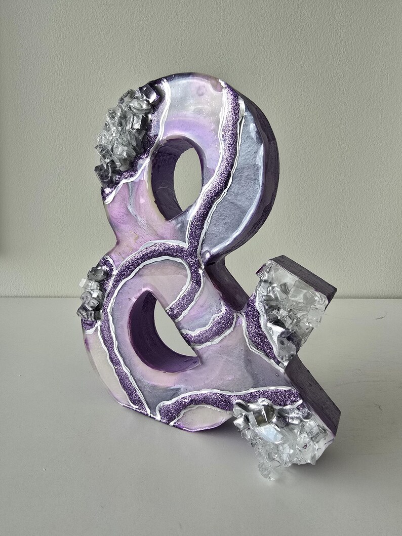 May include: A decorative ampersand sculpture in shades of purple, white, and silver. The sculpture features a glittery, marbled design with crystal-like accents. The ampersand is approximately 8 inches tall.