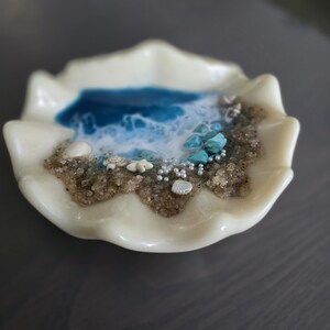 May include: An off-white, seashell-shaped dish featuring a miniature beach scene. The scene includes blue resin representing the ocean, sand, small shells, turquoise stones, and tiny pearl-like beads. The dish has a wavy edge.