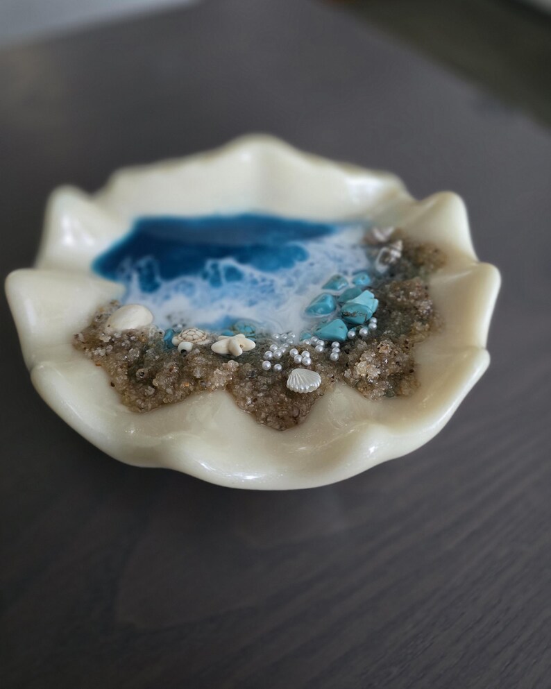 May include: An off-white, seashell-shaped dish featuring a miniature beach scene. The scene includes blue resin representing the ocean, sand, small shells, turquoise stones, and tiny pearl-like beads. The dish has a wavy edge.