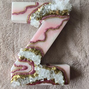 May include: A decorative letter "Z" in pink and white, adorned with gold glitter accents and clear crystal embellishments. The letter has a geode-like design, with a cluster of crystals at the top.