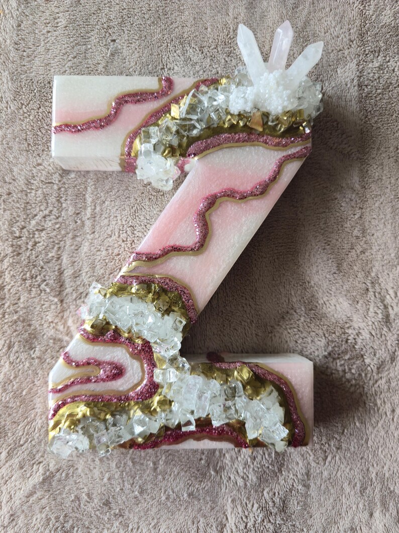 May include: A decorative letter "Z" in pink and white, adorned with gold glitter accents and clear crystal embellishments. The letter has a geode-like design, with a cluster of crystals at the top.