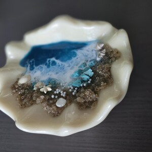 May include: A decorative, off-white seashell-shaped dish with a miniature ocean scene. The scene includes a blue and white wave design, sand, turquoise stones, tiny shells, and pearl-like beads. The dish has a glossy finish.