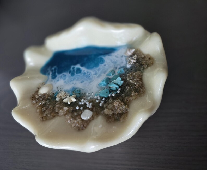 May include: A decorative, off-white seashell-shaped dish with a miniature ocean scene. The scene includes a blue and white wave design, sand, turquoise stones, tiny shells, and pearl-like beads. The dish has a glossy finish.