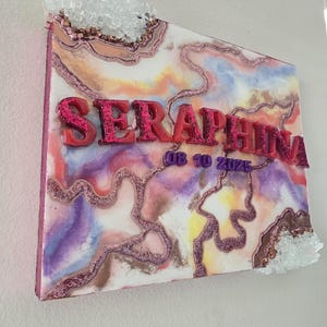 Puede incluir: Una pieza de arte mural decorativa con un dise&ntilde;o abstracto y colorido. La obra presenta el nombre "SERAPHINA" en letras en relieve y brillantes, junto con la fecha "08 10 2025". Racimos de cristales transparentes adornan los bordes superior e inferior.