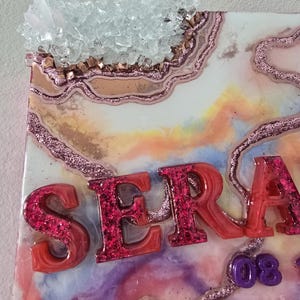 Puede incluir: Una pieza de arte decorativa de resina con el nombre "SERA" en letras rojas con purpurina. La obra presenta un dise&ntilde;o tipo geoda con detalles de purpurina rosa, un grupo de cristales y un fondo colorido y veteado.