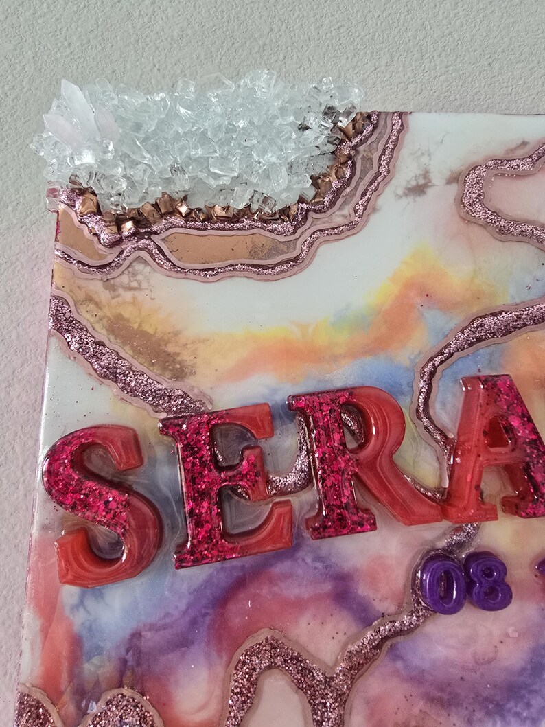 Puede incluir: Una pieza de arte decorativa de resina con el nombre "SERA" en letras rojas con purpurina. La obra presenta un dise&ntilde;o tipo geoda con detalles de purpurina rosa, un grupo de cristales y un fondo colorido y veteado.