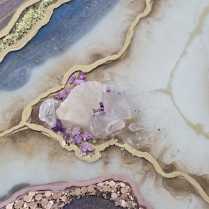 Może przedstawiać: Zbliżenie dekoracyjnego dzieła sztuki z żywicy o wzorze przypominającym geodę. Dzieło sztuki charakteryzuje się odcieniami niebieskiego, fioletowego i białego, zaakcentowanymi złotym brokatem i złotymi liniami. Na powierzchni ułożone są przezroczyste i fioletowe kryształy.