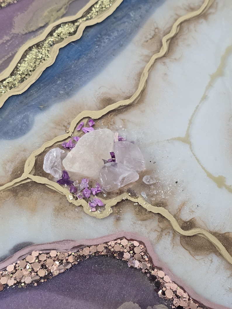 Może przedstawiać: Zbliżenie dekoracyjnego dzieła sztuki z żywicy o wzorze przypominającym geodę. Dzieło sztuki charakteryzuje się odcieniami niebieskiego, fioletowego i białego, zaakcentowanymi złotym brokatem i złotymi liniami. Na powierzchni ułożone są przezroczyste i fioletowe kryształy.