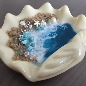 May include: A clam shell-shaped decorative dish with a coastal theme. It features a sandy beach area with turquoise stones, seashells, and a starfish. Blue and white resin simulates ocean waves. The shell is a pearlescent off-white colour.
