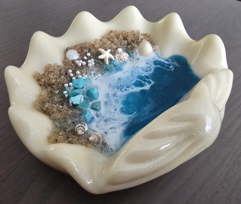 May include: A clam shell-shaped decorative dish with a coastal theme. It features a sandy beach area with turquoise stones, seashells, and a starfish. Blue and white resin simulates ocean waves. The shell is a pearlescent off-white colour.