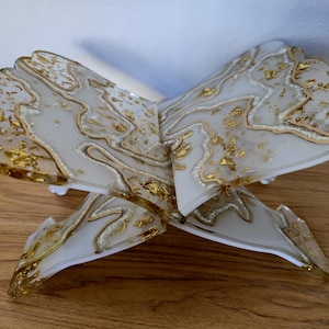 K&ouml;nnte beinhalten: Ein dekorativer Buchst&auml;nder aus klarem Harz mit einem wei&szlig;en und goldenen Marmormuster. Der St&auml;nder ist offen, mit einem gewellten Rand und Goldblattakzenten. Er ist f&uuml;r die Aufnahme eines Buches oder anderer Gegenst&auml;nde konzipiert.