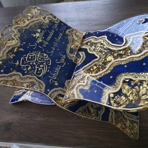 May include: A decorative book stand with a blue and gold design. The stand features intricate gold leaf accents and calligraphy on a blue background. The text reads "So remember Me, I will remember You, Surah Al-Baqarah 2:152, Arhama Syed."