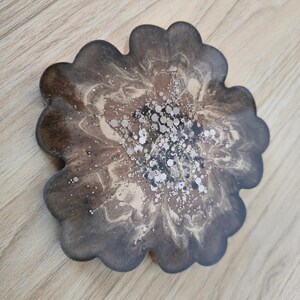 K&ouml;nnte beinhalten: Ein bl&uuml;tenf&ouml;rmiger Untersetzer mit einem dunkelbraunen, beigen und silbernen Design. Der Untersetzer hat einen gewellten Rand und ein marmoriertes Aussehen. Die Mitte des Untersetzers hat silberne Glitzerakzente.