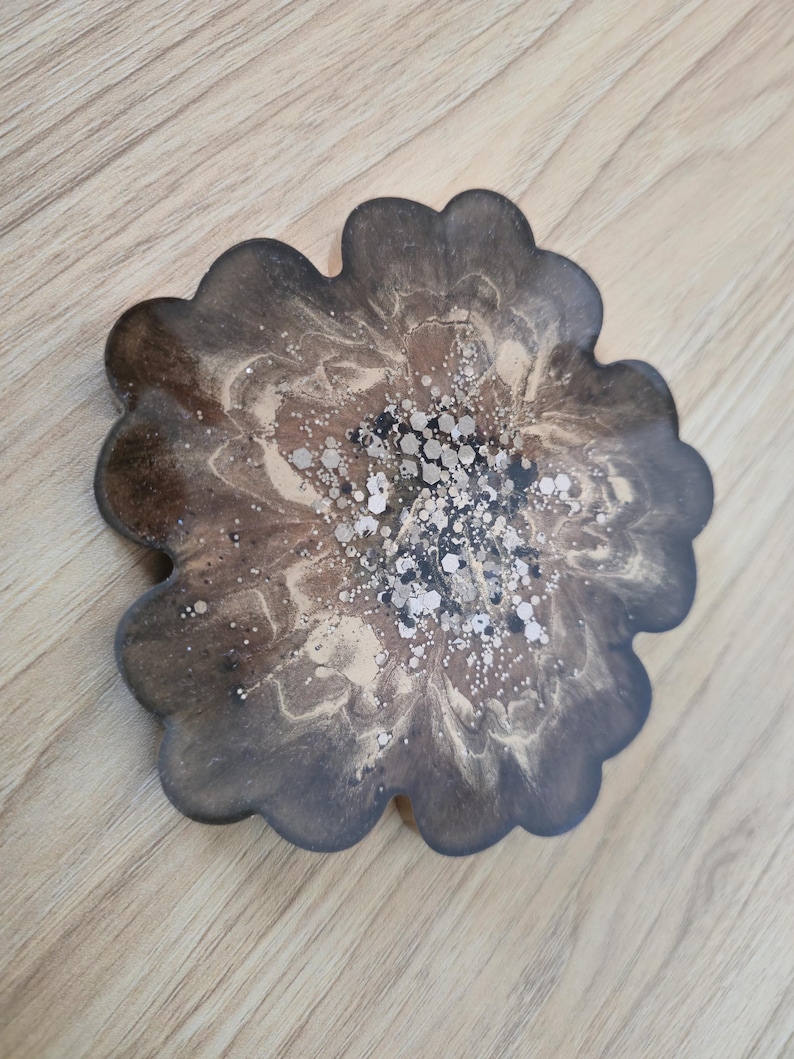 K&ouml;nnte beinhalten: Ein bl&uuml;tenf&ouml;rmiger Untersetzer mit einem dunkelbraunen, beigen und silbernen Design. Der Untersetzer hat einen gewellten Rand und ein marmoriertes Aussehen. Die Mitte des Untersetzers hat silberne Glitzerakzente.