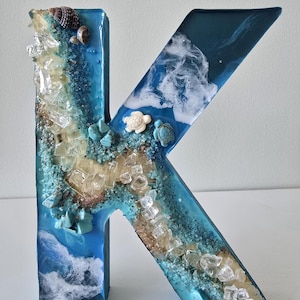 Może przedstawiać: Dekoracyjna litera "K" wykonana z żywicy, z motywem plażowym. Litera jest gł&oacute;wnie niebieska z wbudowanymi elementami, takimi jak muszle, piasek, przezroczyste kryształy i małe figurki ż&oacute;łwi. Projekt nawiązuje do tematu oceanu lub wybrzeża.