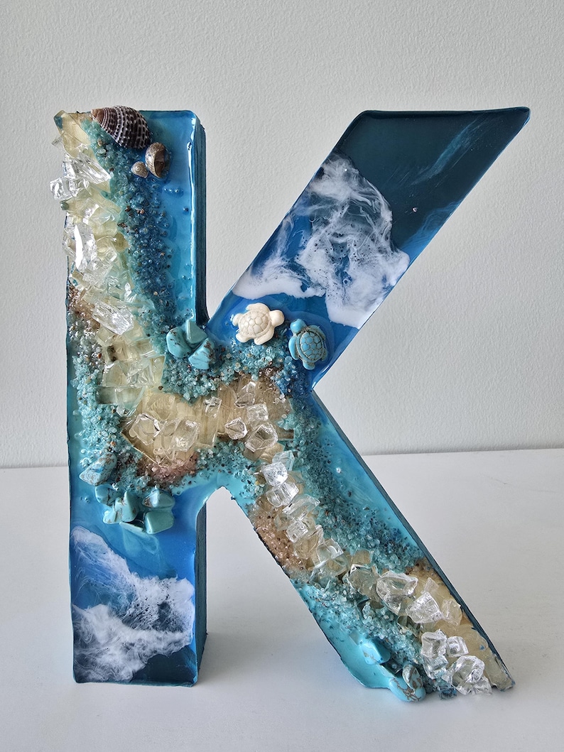 Może przedstawiać: Dekoracyjna litera "K" wykonana z żywicy, z motywem plażowym. Litera jest gł&oacute;wnie niebieska z wbudowanymi elementami, takimi jak muszle, piasek, przezroczyste kryształy i małe figurki ż&oacute;łwi. Projekt nawiązuje do tematu oceanu lub wybrzeża.