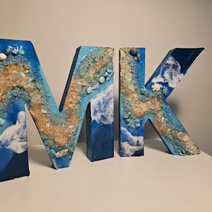 Może przedstawiać: Dekoracyjne drewniane litery tworzące napis "MK", pomalowane w odcieniach niebieskiego, beżu i bieli, z zatopionymi muszlami i kawałkami szkła. Litery mają motyw nadmorski, przypominający scenę na plaży.