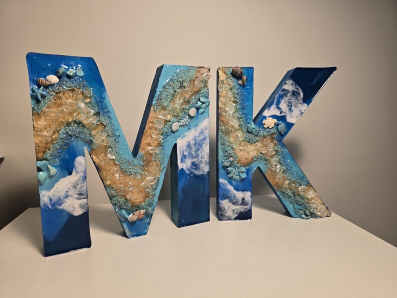 Może przedstawiać: Dekoracyjne drewniane litery tworzące napis "MK", pomalowane w odcieniach niebieskiego, beżu i bieli, z zatopionymi muszlami i kawałkami szkła. Litery mają motyw nadmorski, przypominający scenę na plaży.