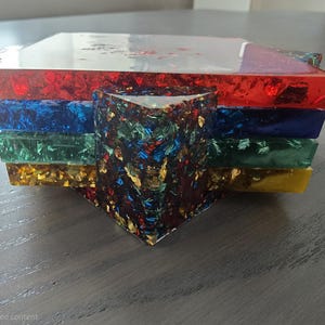Pode incluir: Um conjunto de bases para copos de resina coloridas. As bases para copos s&atilde;o vermelhas, azuis, verdes e amarelas, com uma camada superior transparente. A pe&ccedil;a central &eacute; um prisma triangular multicolorido. Feito com flocos de folha de ouro.