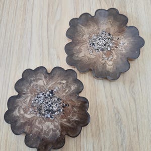 K&ouml;nnte beinhalten: Zwei bl&uuml;tenf&ouml;rmige Untersetzer mit dunkelbraunem, marmoriertem Design. Die Mitte jedes Untersetzers ist mit silbernem und schwarzem Glitzer gef&uuml;llt. Die Untersetzer sind aus Kunstharz gefertigt und liegen auf einer hellbraunen Holzoberfl&auml;che.