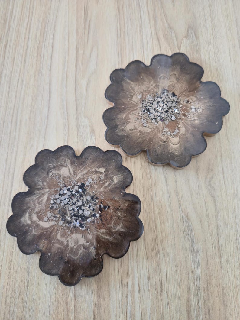 K&ouml;nnte beinhalten: Zwei bl&uuml;tenf&ouml;rmige Untersetzer mit dunkelbraunem, marmoriertem Design. Die Mitte jedes Untersetzers ist mit silbernem und schwarzem Glitzer gef&uuml;llt. Die Untersetzer sind aus Kunstharz gefertigt und liegen auf einer hellbraunen Holzoberfl&auml;che.