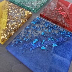 Pode incluir: Um conjunto de cinco bases quadradas de resina em v&aacute;rias cores. As bases apresentam tons dourados, azuis, vermelhos, verdes e amarelos, com detalhes em folha met&aacute;lica embutidos. As bases est&atilde;o dispostas sobre uma superf&iacute;cie cinza.