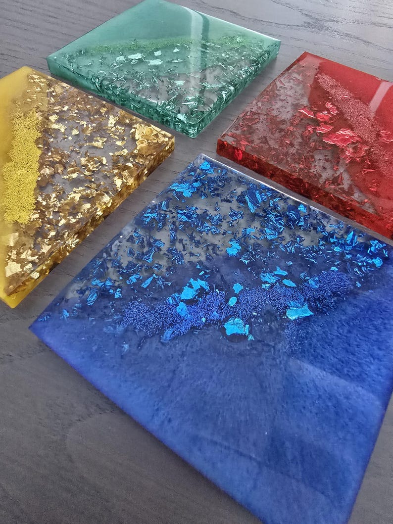 Pode incluir: Um conjunto de cinco bases quadradas de resina em v&aacute;rias cores. As bases apresentam tons dourados, azuis, vermelhos, verdes e amarelos, com detalhes em folha met&aacute;lica embutidos. As bases est&atilde;o dispostas sobre uma superf&iacute;cie cinza.