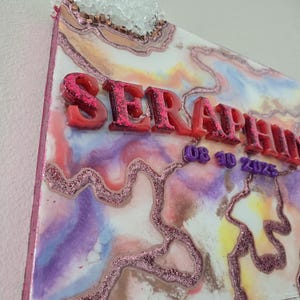 Puede incluir: Una colorida obra de arte abstracta en resina con la palabra "SERAPHINA" en letras rojas con purpurina. La obra presenta una mezcla de tonos rosas, morados, azules y amarillos, detalles con purpurina y un grupo de cristales en la parte superior.