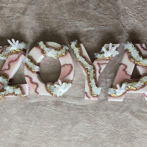 May include: The name "ZOYA" is spelled out in decorative letters. Each letter is white with pink and gold accents, and embellished with clear crystal clusters. The letters are arranged on a neutral-colored surface.