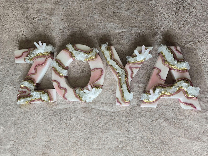 May include: The name "ZOYA" is spelled out in decorative letters. Each letter is white with pink and gold accents, and embellished with clear crystal clusters. The letters are arranged on a neutral-colored surface.