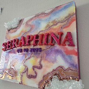 Puede incluir: Una pieza de arte mural decorativa con el nombre "SERAPHINA" en letras rosas brillantes. La obra de arte presenta un dise&ntilde;o abstracto colorido con acentos rosas, morados y dorados, y adornos de cristal.