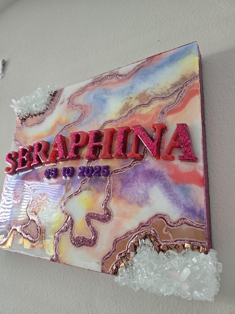Puede incluir: Una pieza de arte mural decorativa con el nombre "SERAPHINA" en letras rosas brillantes. La obra de arte presenta un dise&ntilde;o abstracto colorido con acentos rosas, morados y dorados, y adornos de cristal.
