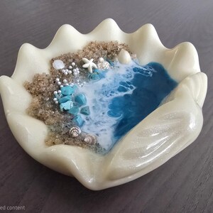 May include: A decorative, off-white clam shell-shaped dish with a coastal scene. The interior features a blue ocean, sandy beach, seashells, turquoise stones, and a starfish. The dish is a unique home decor item.